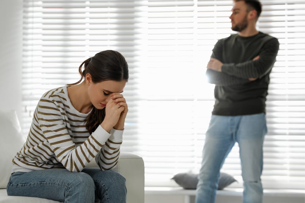 Nainen istuu sohvalla pää painuneena ja kädet yhteen puristettuina, taustalla mies seisoo ikkunan edessä kädet ristissä, huoneessa vallitsee etäinen ja ristiriitainen tunnelma.