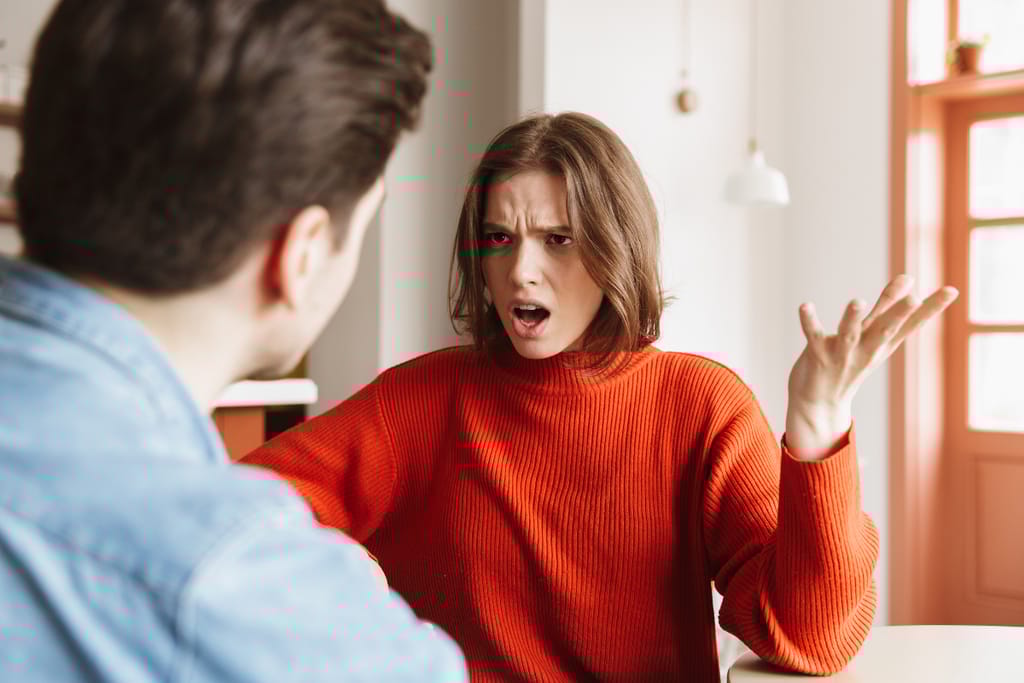 Kuvassa nainen punainen villapaita päällään raivoaa miehelle. Nainen on vihainen ja levittelee käsiään