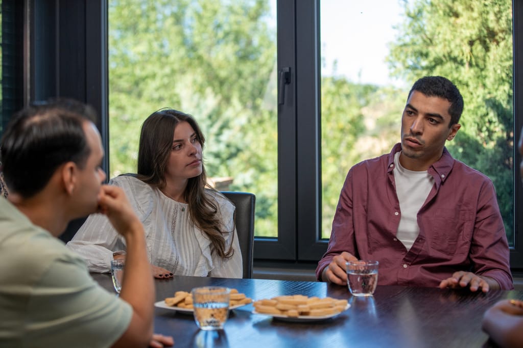 Kolme henkilöä istuu ruokapöydän ääressä kirkkaassa päivänvalossa. Nainen valkoisessa paidassa ja mies vaaleanpunaisessa kauluspaidassa kuuntelevat vakavina, kun kolmas henkilö puhuu. Pöydällä on laseja ja keksejä, ja ilmapiiri vaikuttaa jännittyneeltä ja keskittyneeltä.