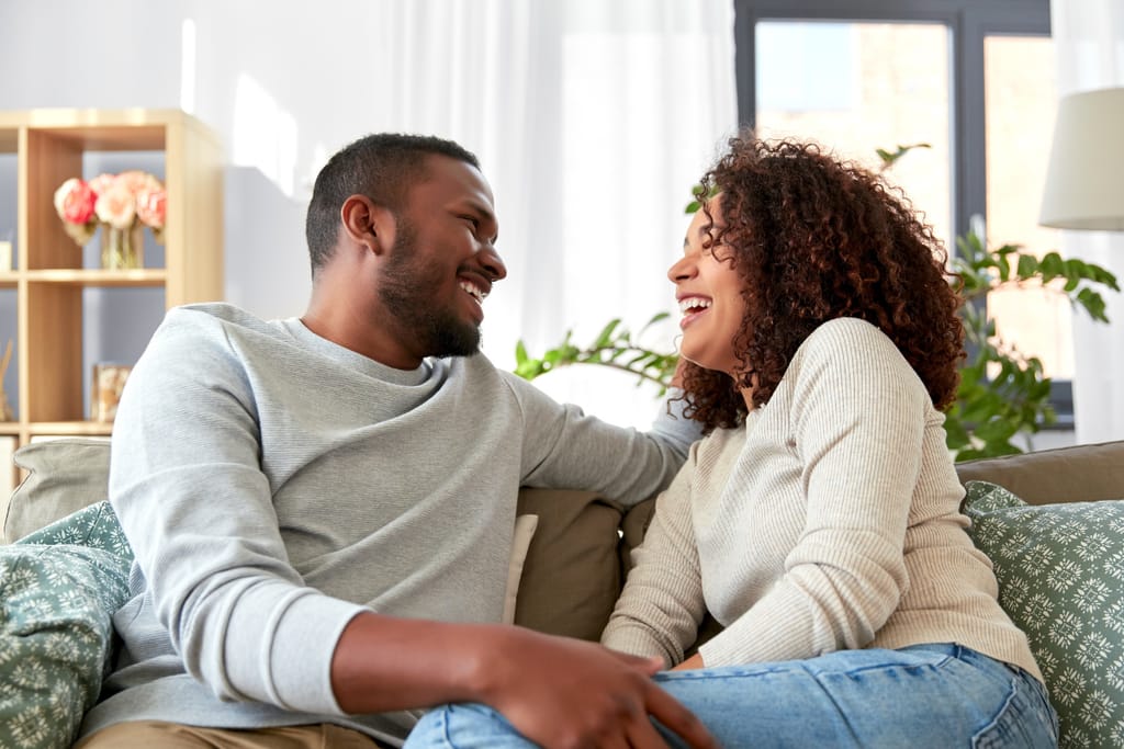 Pariskunta istuu sohvalla valoisassa olohuoneessa, nauraa ja katsoo toisiaan iloisesti. Takana näkyy viherkasveja ja kirjahylly, ja huoneen sisustus luo kodikkaan ja läheisen tunnelman.