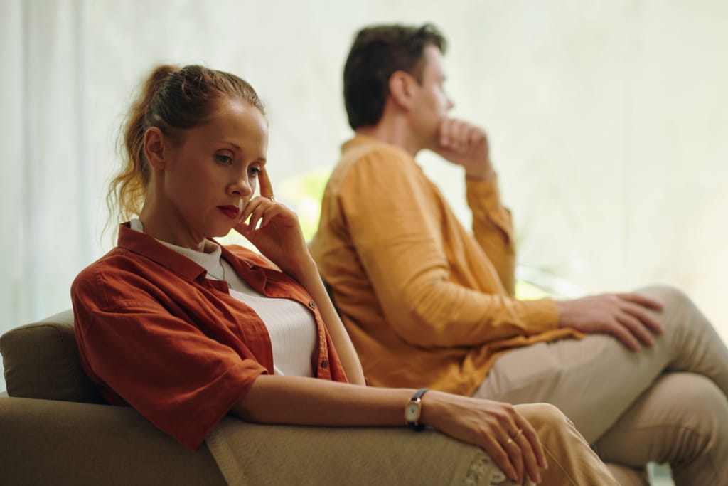 Nainen ja mies istuvat sohvalla selin toisiinsa, molemmat mietteliään ja etäisen näköisinä. Nainen katsoo alas käsi poskellaan, mies istuu taustalla nojaten käteensä ja katselee poispäin. Kuvassa on jännittynyt ja hiljainen tunnelma, joka viestii parisuhteen ristiriidasta tai vieraantumisesta.