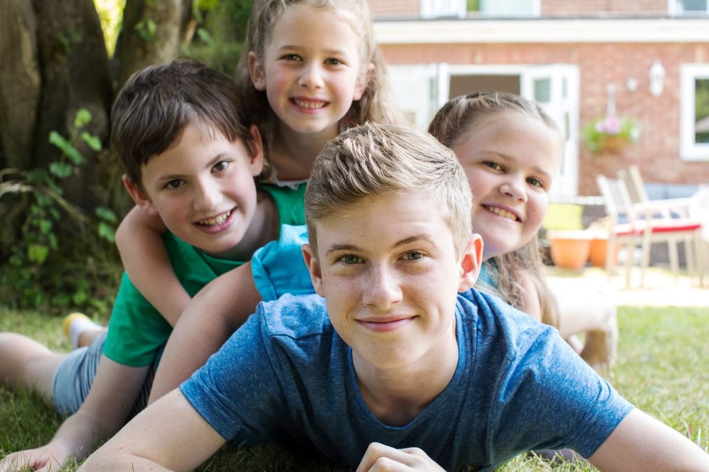 Sisarukset makaavat nurmikolla jonossa, jossa isoveli on etummaisena ja nuoremmat perässä kokojärjestyksessä. He hymyilevät kameralle. 