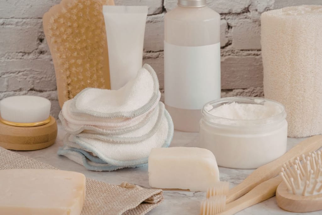 Erilaisia kylpyhuoneen ja ihonhoidon tuotteita kivipinnalla. Kuvassa näkyy saippuoita, pesulappuja, pulloja, purkki ja puisia harjoja vaaleaa tiiliseinää vasten. 