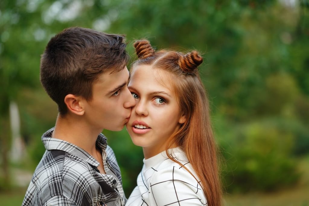 Teini-ikäinen seurustelupari. Poika pussaa tyttöä poskelle, ja tyttö katsoo poispäin pojasta ja kamerasta. He ovat kesäisessä ympäristössä. 