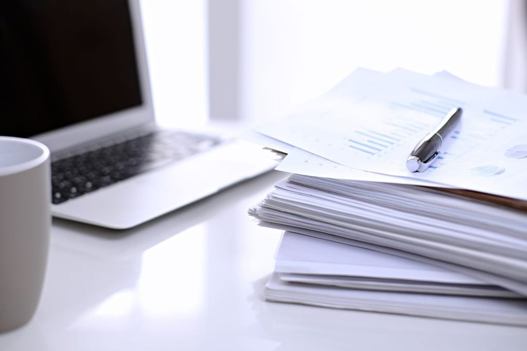 Läppäri, paperipino ja kahvikuppi valkoista taustaa vasten työpisteellä.