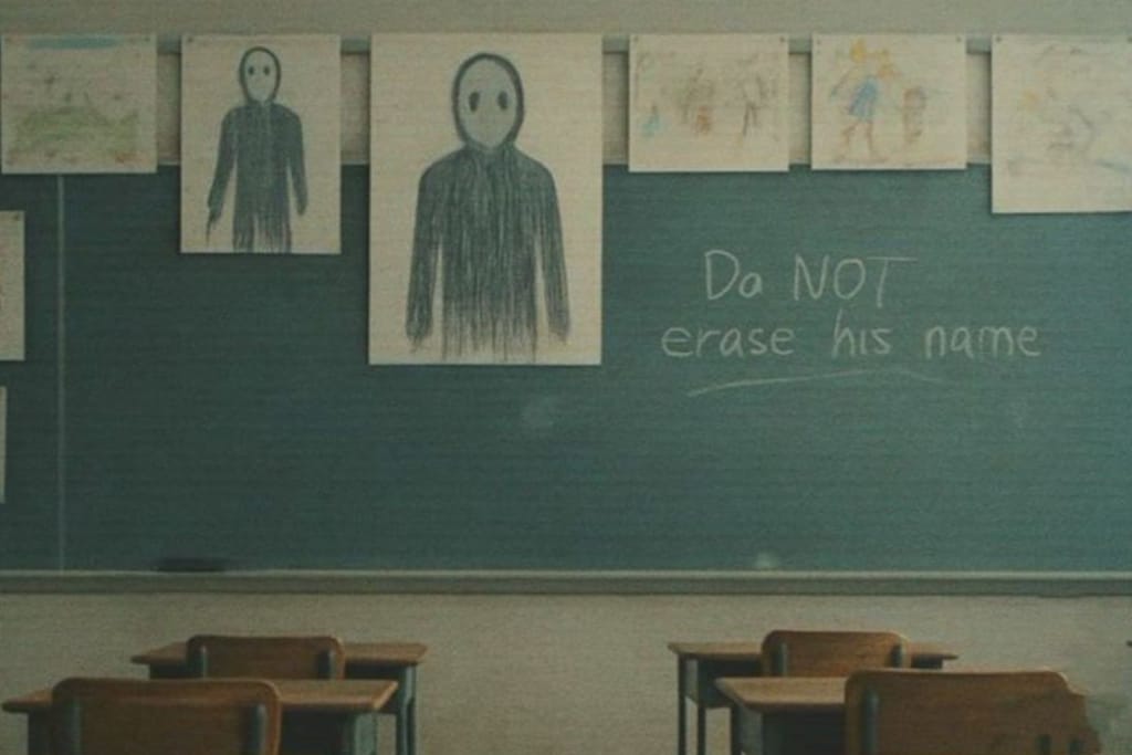 Luokkahuoneen liitutaululle on ripustettu useita lasten piirustuksia, joissa toistuu sama tummaan sävyyn piirretty, suuton ihmishahmo. Liitutaulussa näkyy käsin kirjoitettu teksti: “Do NOT erase his name”. Etualalla on tyhjiä koulupulpetteja.