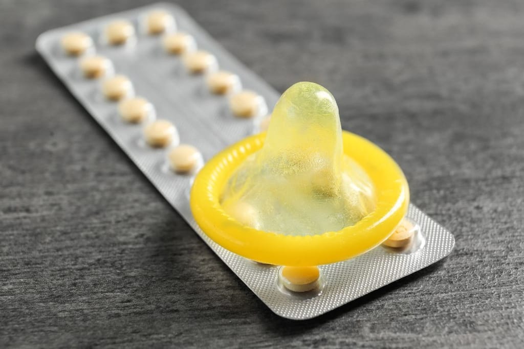 E-pilleriliuska ja keltainen kondomi päällekäin harmaata taustaa vasten. 