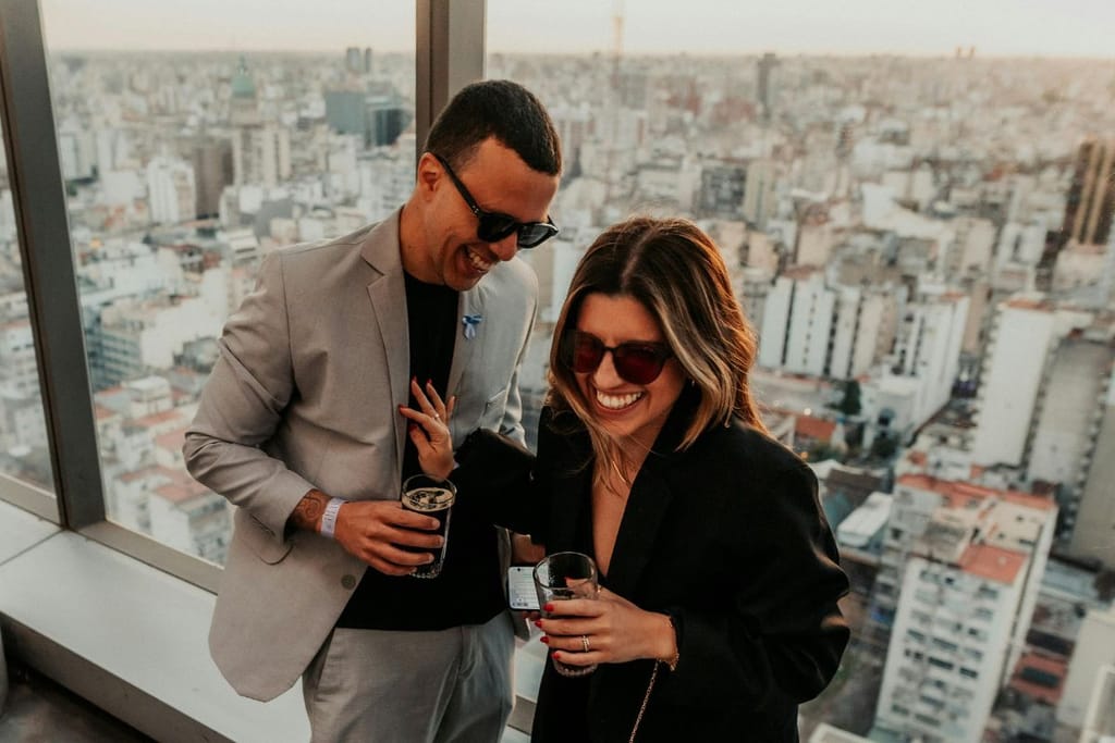 Hymyilevä pari nauraa toisilleen kaupungin kattoterassilla. Alla avautuu iso miljoonakaupunki. Molemmilla on juomat kädessä ja heillä on hauskaa, nainen nauraa niin, että hampaat vilkkuvat. 