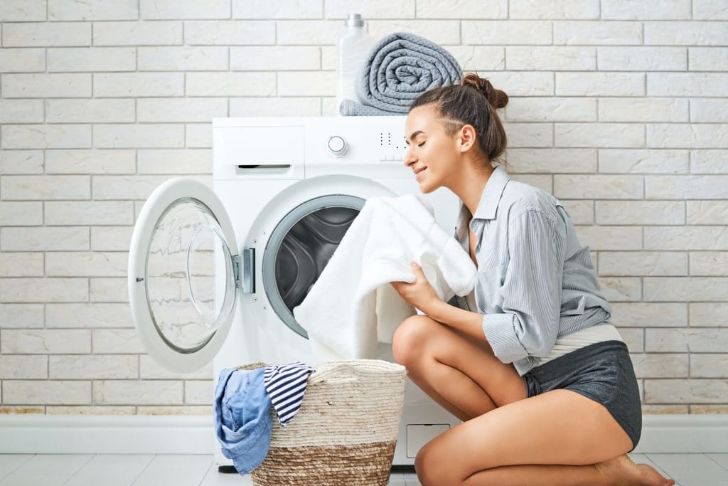 Nainen haistelee puhdasta pyyhettä tyytyväisen näköisenä pyykkikoneen edessä.
