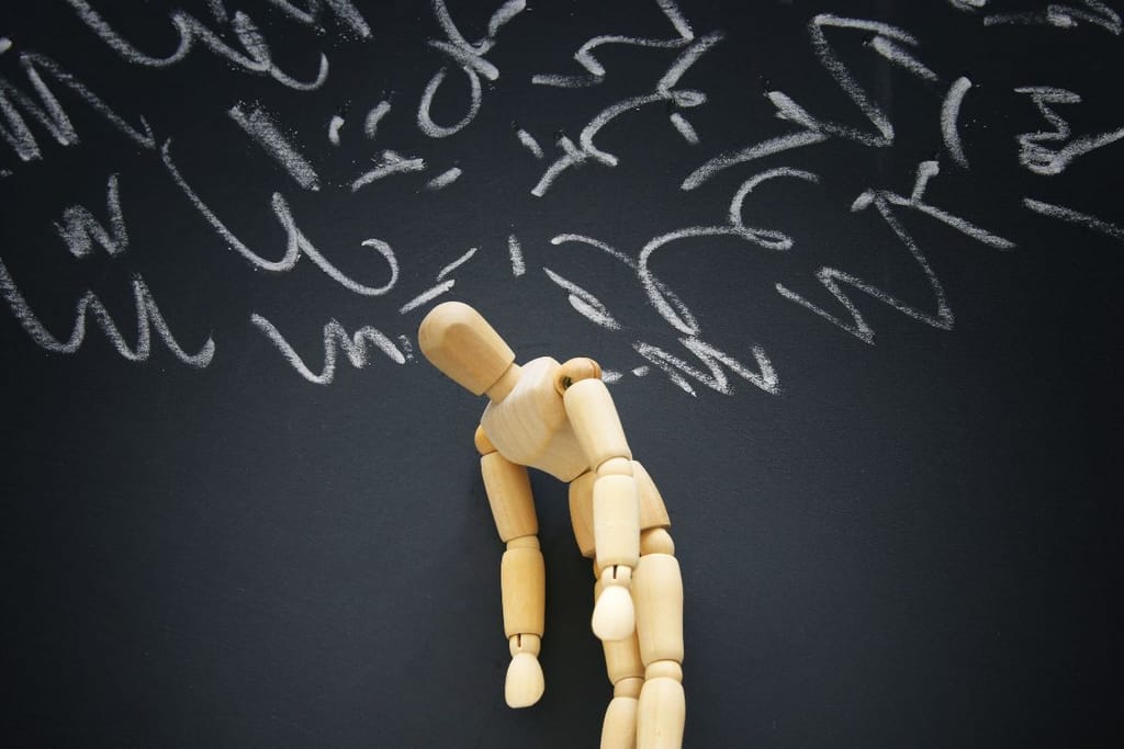 Puinen mallinukke kumartunut liitutaulun edessä. Sen pään yläpuolelle on piirretty ajatuskuplaksi koukeroita, jotka kuvaavat mielen sekavuutta.