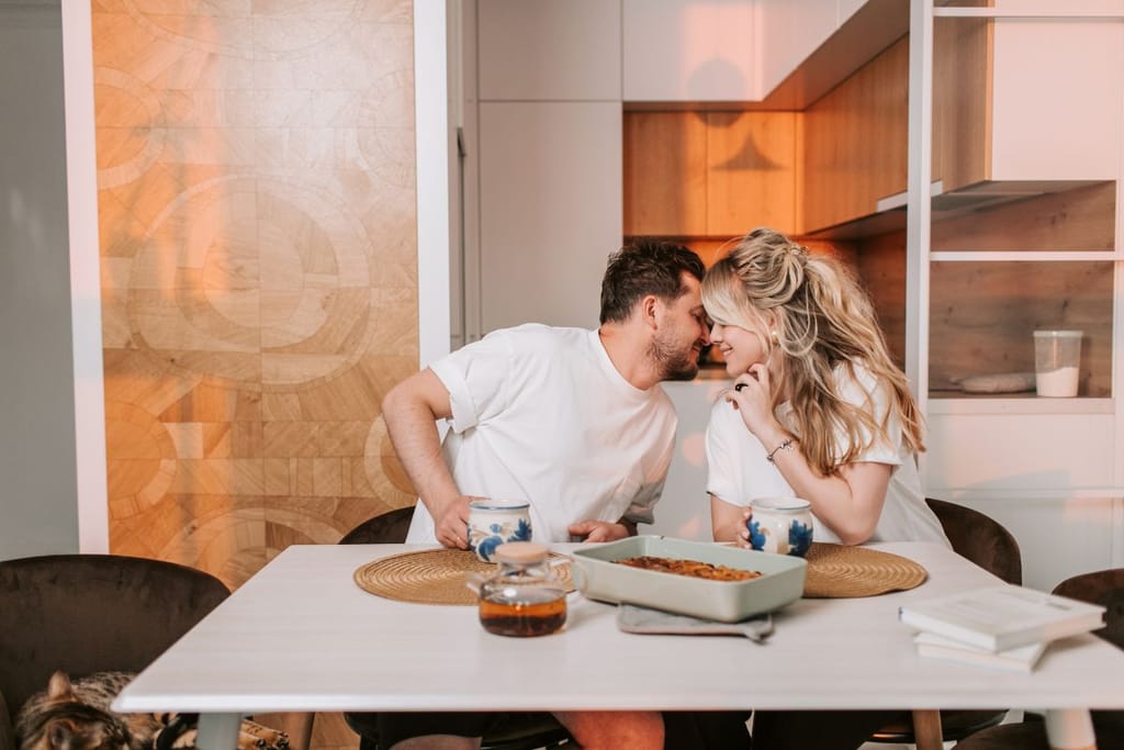 Pariskunta istuu keittiön pöydän ääressä vastakkain, kahvikupit edessään. He nojaavat toisiaan kohti ja koskettavat otsiaan.