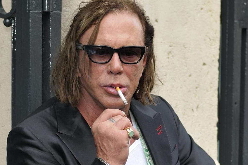 Mickey Rourke mustissa aurinkolaseissa ja tummassa puvuntakissa tupakka huulillaan, kuvattuna ulkona vaaleaa seinää vasten.