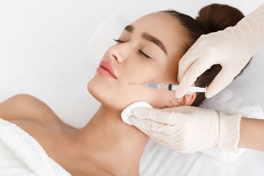 Henkilö makaa hoitopöydällä, ja toinen henkilö antaa pistosta kasvoihin suojakäsineet kädessä.
