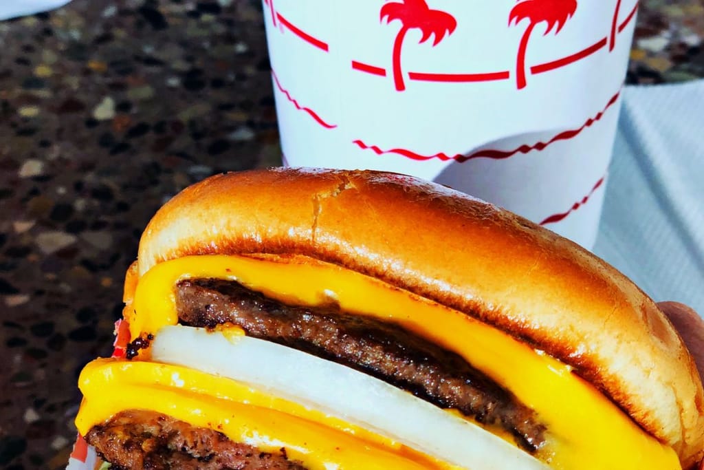 Lähikuva In-N-Out-hampurilaisesta, jossa on sämpylä, kaksi pihviä, sulanut juusto ja sipulia. Taustalla näkyy pikaruokaketjun logolla kuvioitu juomamuki.