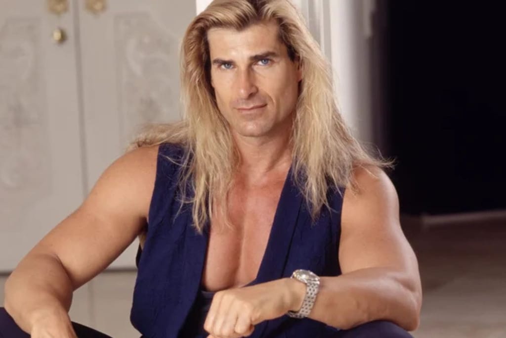 Kuvassa on Fabio Lanzoni, 1990-luvun tunnettu romanssikirjojen kansimalli ja popkulttuuri-ikoni. Hän katsoo suoraan kameraan vakavana, pitkät vaaleat hiukset hartioille laskeutuen ja yllään tummansininen liivi. Kuva henkii tyypillistä 90-luvun glamouria ja Fabion itsevarmaa karismaa.