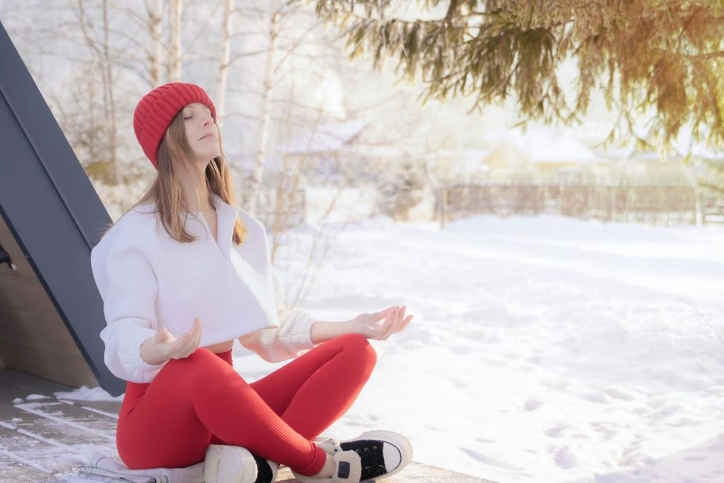 Nainen istuu ulkona lumisessa ympäristössä jalat ristissä ja meditoi rauhallisesti silmät suljettuina.