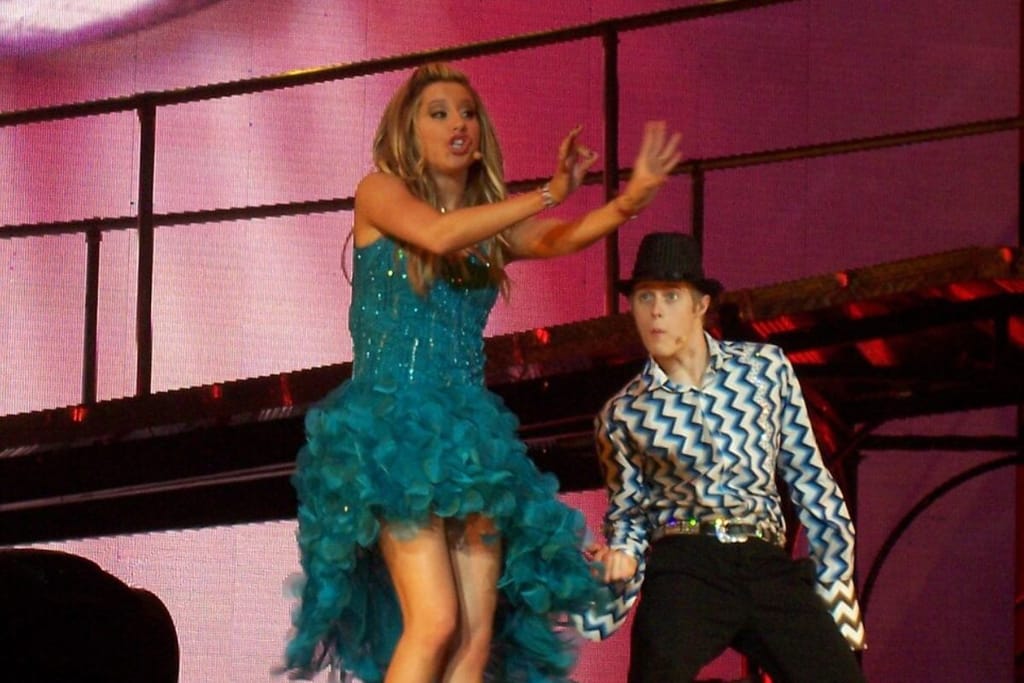 Ashley Tisdale esiintyy turkoosissa, röyhelöisessä mekossa laulaa tai puhuu lavalla kädet koholla, vieressään tanssija Lucas Grabeel kuvioidussa paidassa ja hatussa värikkään lavataustan edessä.