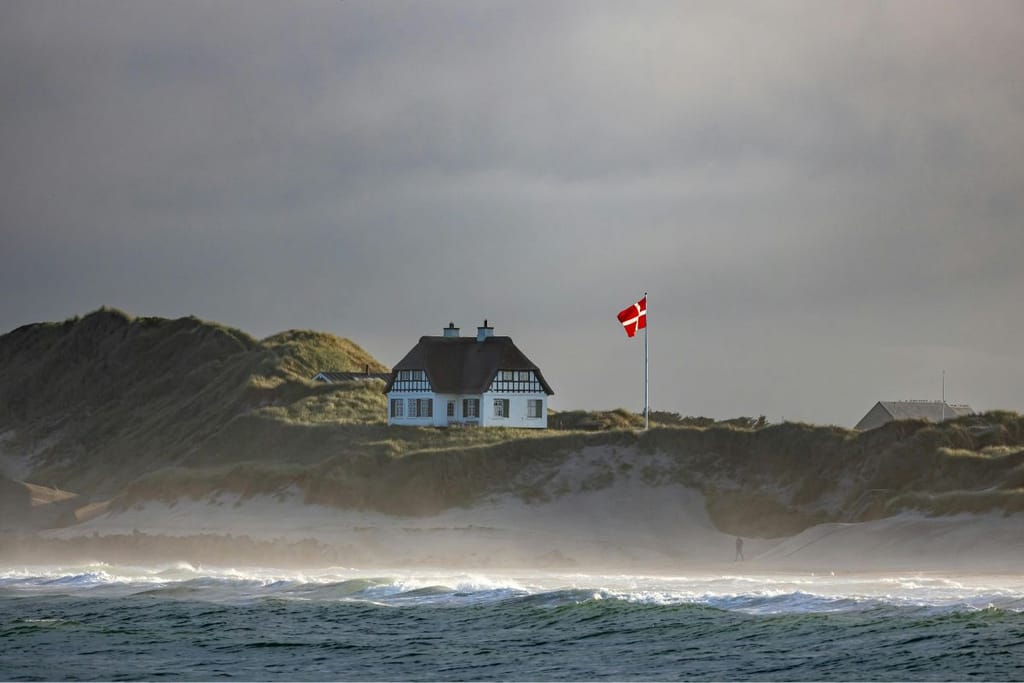 Rantadyynien päällä sijaitseva vaalea talo, jonka vieressä liehuu Tanskan lippu. Etualalla näkyy aaltoileva meri ja taustalla harmaa, pilvinen taivas.