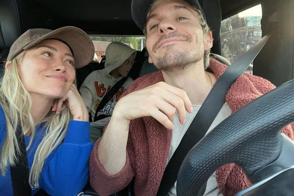 Hilary Duff ja hänen aviomiehensä autossa. Molemmilla on turvavyöt ja takapenkillä istuu huppupäinen lapsi. Hilary katsoo miestään ja mies katsoo kameran ohi ja lisäksi he näyttävät iloisilta. 