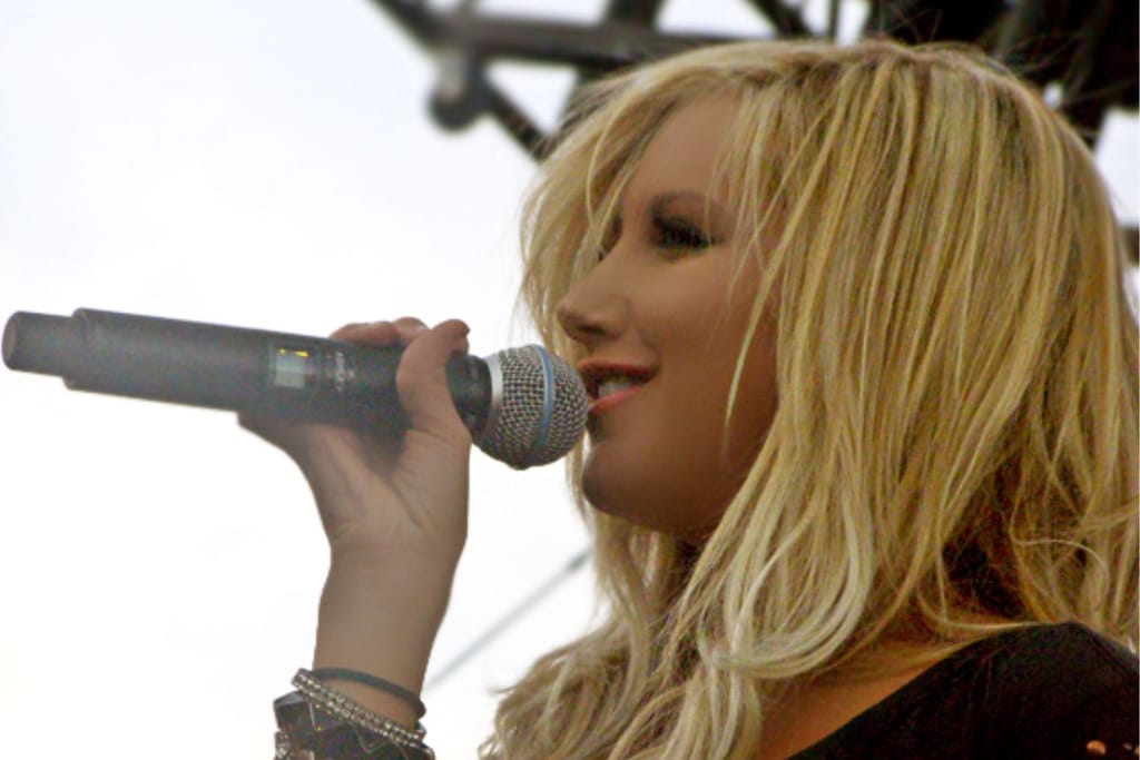 Ashley Tisdale laulaa mikrofoniin. Hän on taustarakenteista päätellen lavalla ja taustalla näkyy kirkas taivas. 