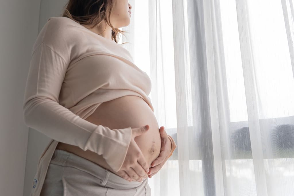 Raskaana oleva nainen seisoo ikkunan vieressä ja pitää käsiään vatsansa päällä. 