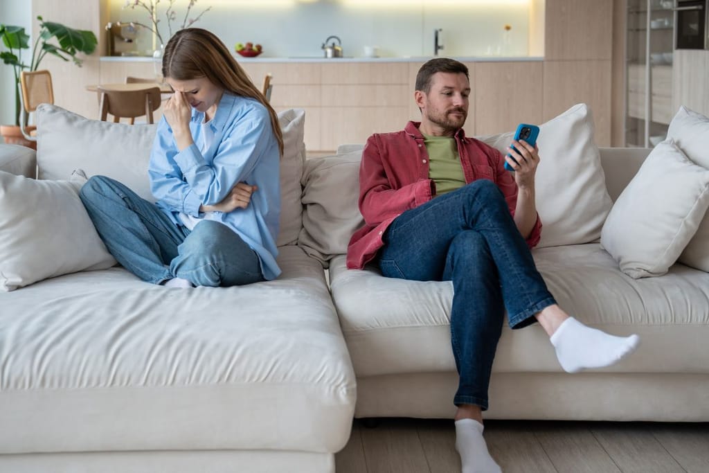 Nainen istuu sohvalla selvästi ahdistuneena, vieressä oleva mies keskittyy älypuhelimeensa.