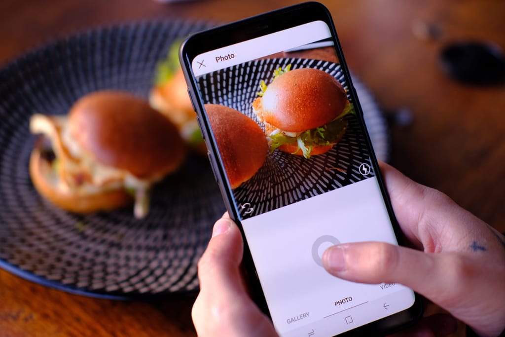 Ihminen ottaa älypuhelimella kuvaa kahdesta hampurilaisesta sosiaaliseen mediaan.