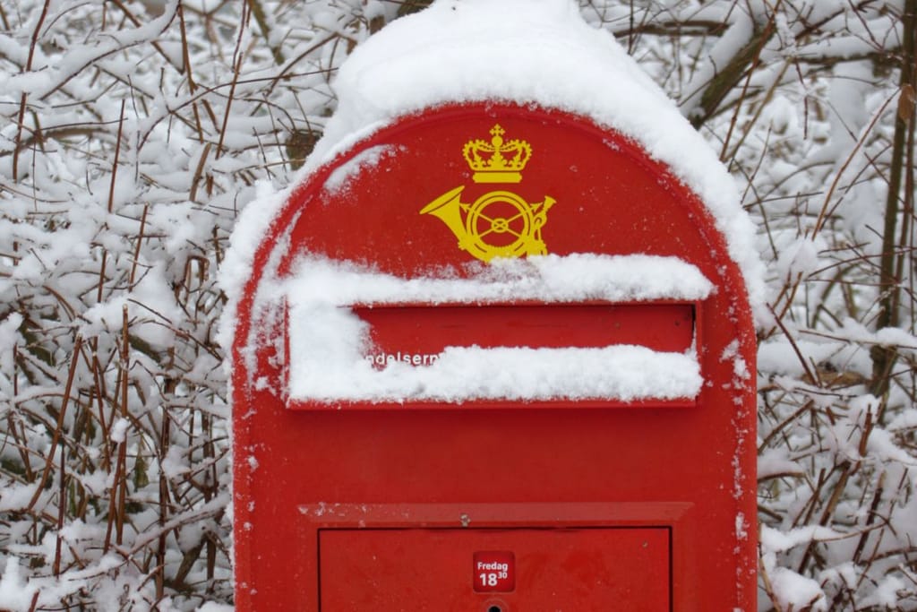 Tanskalainen postilaatikko, jossa on postin logo. Laatikko on peittynyt lumeen. 