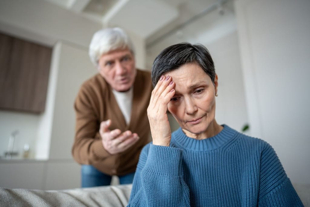Iäkäs mies elehtii taustalla, kun etualalla oleva nainen pitää kättään otsallaan ja näyttää uupuneelta ja huolestuneelta.