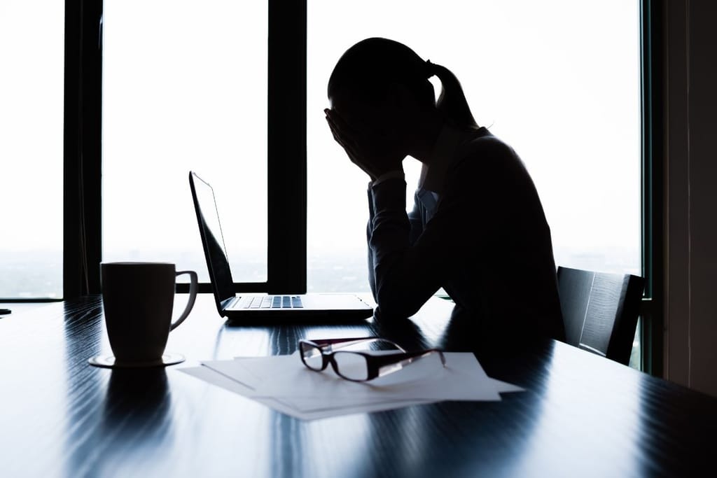 Henkilö istuu tietokoneen ääressä toimistossa ja peittää kasvonsa käsillään, selvästi stressaantuneena.