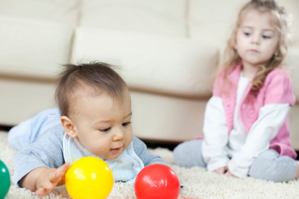 Vauva leikkii lattialla värikkäillä palloilla, kun taustalla istuva vanhempi sisarus seuraa tilannetta vakavana.
