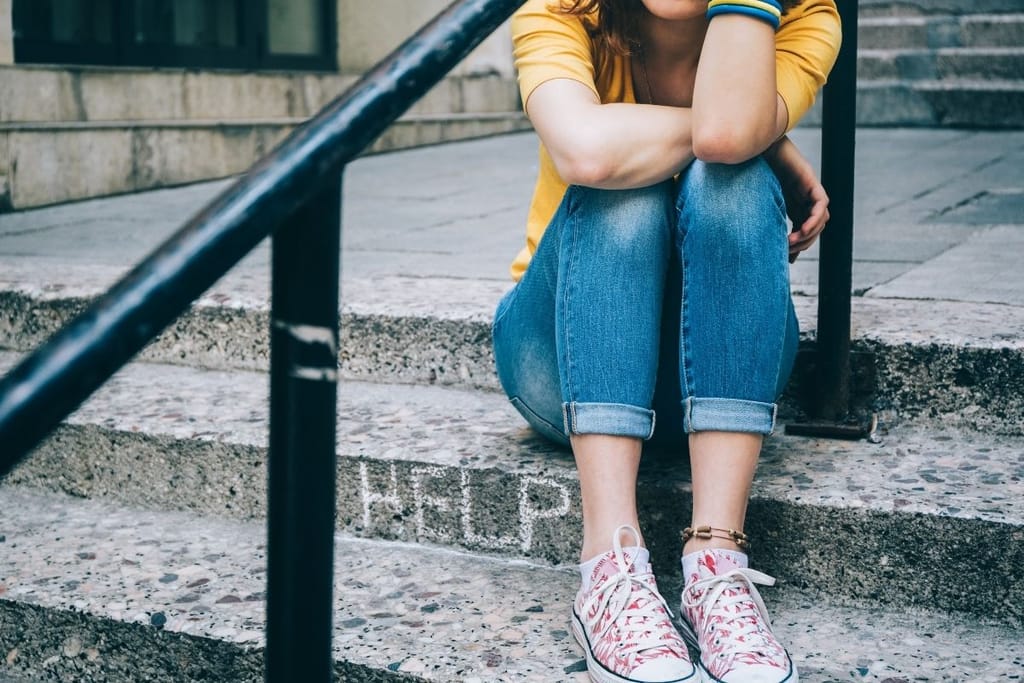 Nuori istuu portailla mustan metallikaiteen vieressä, johon on kirjoitettu liidulla HELP. Kasvoja ei näy.