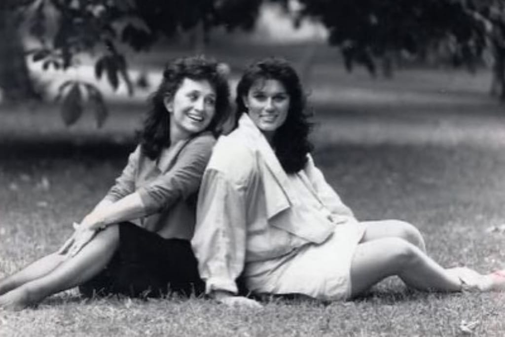 Lucy yhdessä Desirée tyttärensä kanssa nurmikolla. Kuva on mustavalkoinen otos ja naiset poseeraavat selät vastakkain. Äiti katsoo tytärtään ja tytär kameraan ja hymyilee leveästi. 