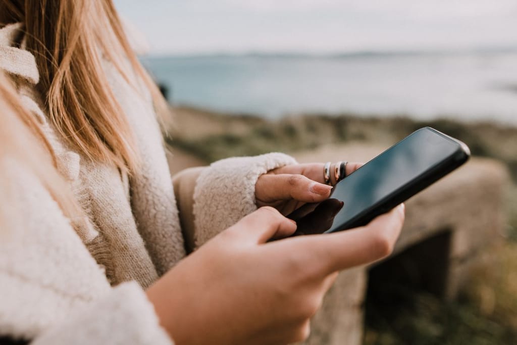 Nainen seisoo ulkona meren äärellä ja kirjoittelee puhelimella. Hänellä on peukalossa sormus ja valkoinen teddykarvainen takki.