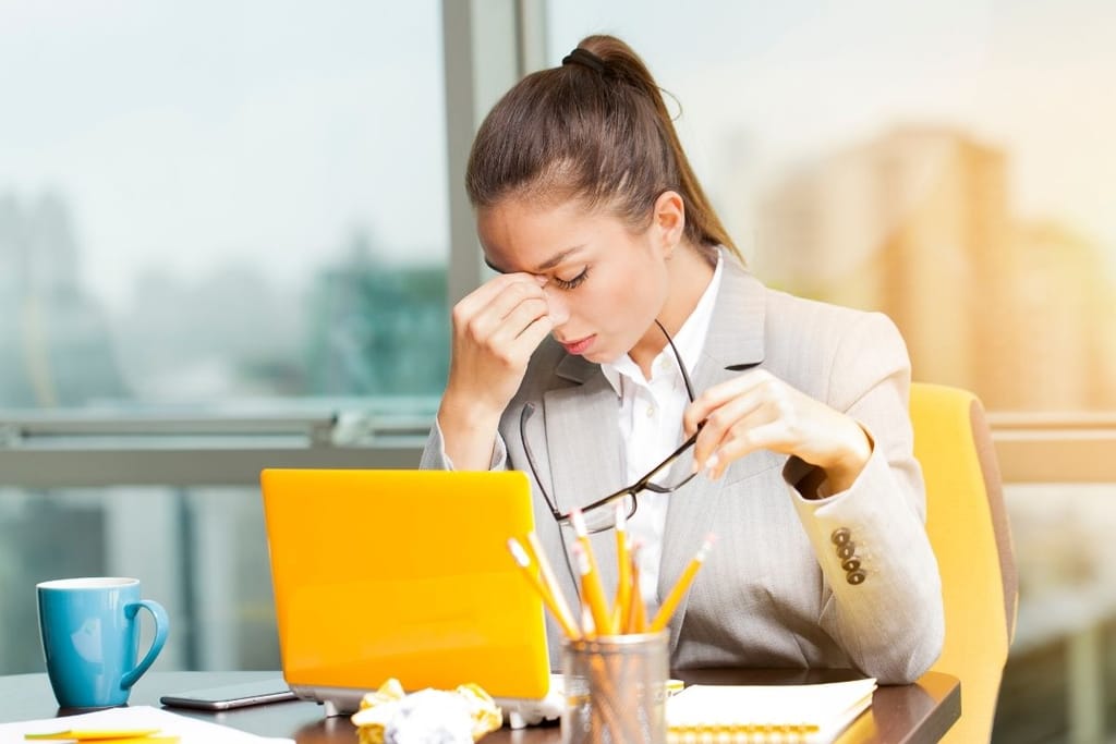 Nainen istuu työpöydän ääressä ja vaikuttaa uupuneelta. Hänellä on kädessään silmälasit ja hän hieroo silmiään.