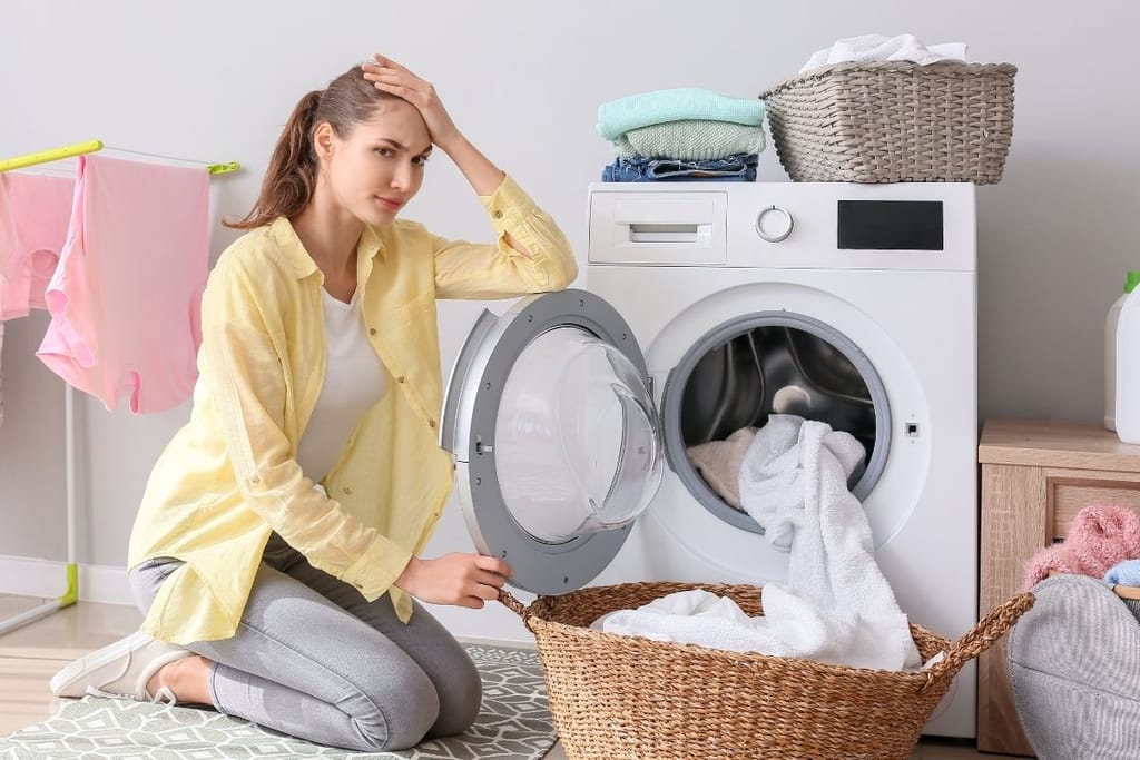 Nainen näyttää tuskastuneelta pyykkien kanssa. Pesukoneen rumpu on auki ja pyykit vedetty osittain pyykkikoriin.