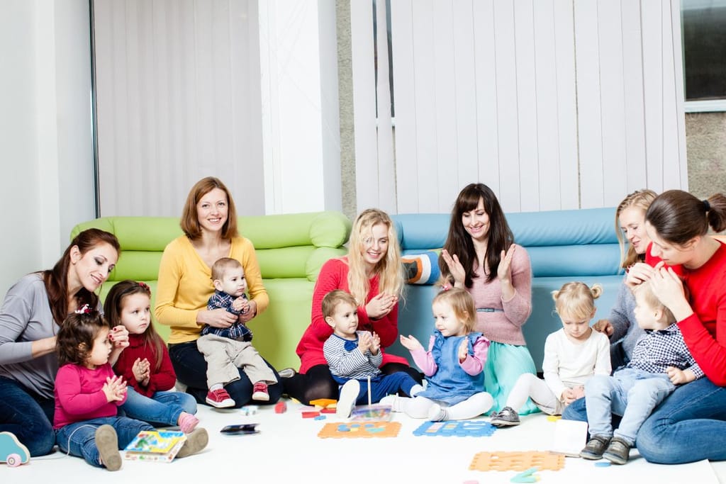 Ryhmä pieniä lapsia ja aikuisia istuu lattialla kirkkaassa leikkitilassa värikkäiden sohvien edessä; osa taputtaa ja lapset leikkivät palikoilla ja leluilla.