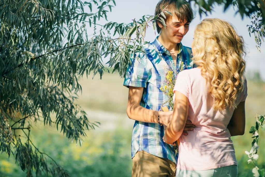Nuoripari seisoo ilta-auringossa ja katsovat toisiaan vastakkain puun juurella. Mies hymyilee. Naisella on pitkät, vaaleat kiharat. He ovat selvästi hullaantuneita. 