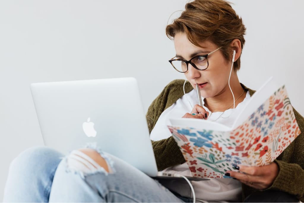 Silmälasipäinen nainen istuu kannettavan tietokoneen ääressä, kuuntelee kuulokkeilla ja selaa samalla muistikirjaa kynä kädessä.