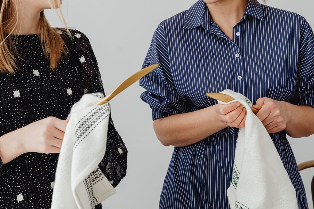 Kaksi henkilöä seisoo vierekkäin ja pitelee käsissään keittiövälineitä, joita he kuivaavat keittiöpyyhkeillä.
