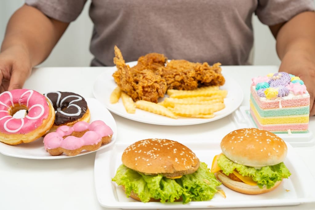 Pöydälle katettu valikoima pikaruokaa ja makeisia: hampurilaisia, ranskalaisia, friteerattua kanaa, donitseja ja värikäs kakkupala. Henkilön kädet näkyvät pöydän ääressä.