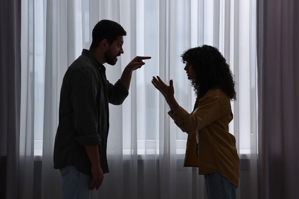 Pariskunta riitelee ikkunan edessä ja osoittelee toisiaan sormillaan. 
