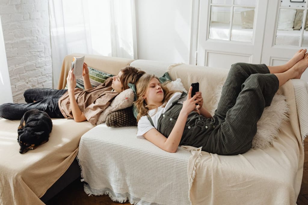 Sisarukset makaavat päät vastakkain sohvalla. Toinen lukee kirjaa ja toinen selailee puhelinta jalat kohotettuna. Valkea tausta, joka vaikuttaisi olohuoneelta. 