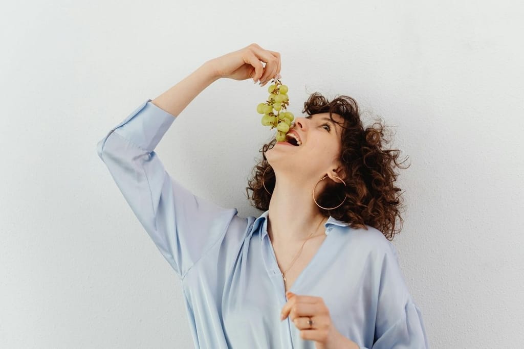 Nainen seisoo vaaleaa taustaa vasten ja on syövinään rypäleitä huvittunut ilme kasvoillaan. Hänellä on kihara tukka ja vaalea paita. 