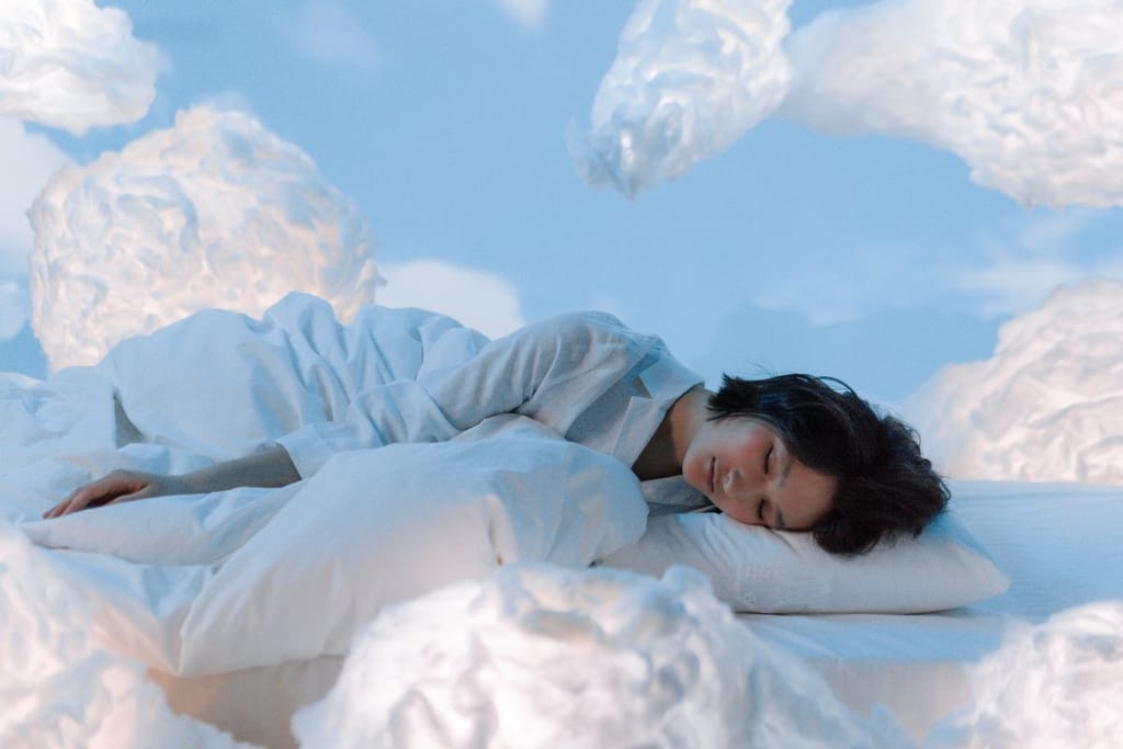 Nainen nukkuu patjalla valkoisissa yövaatteissa pilvien keskellä sinistä taivasta vasten. 