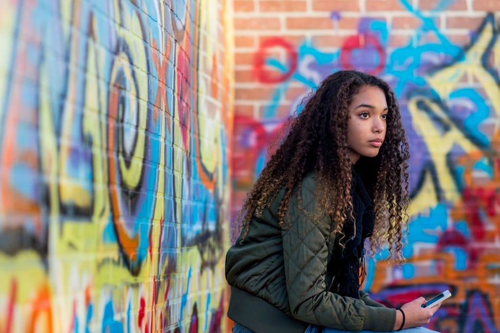 Nuori kiharatukkainen nainen on kuin olisi lukenut jotain todella typerää. Hän istuu graffittiseinän vierellä vihreä takki yllään. 