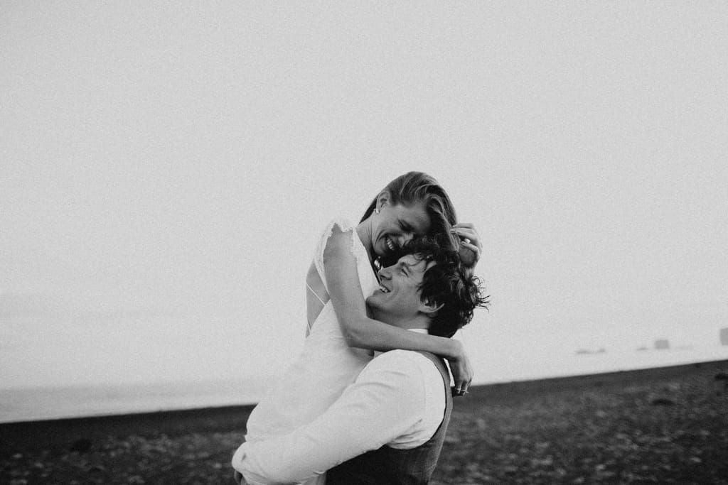 Mustavalkoinen kuva pariskunnasta rannalla. Toinen nostaa toista syliinsä, ja he nauravat.