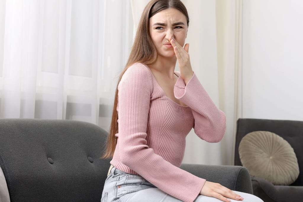 Nuori nainen istuu sohvalla vaaleassa huoneessa ja peittää nenänsä kädellään. Hän näyttää irvistävän epämiellyttävälle hajulle.
