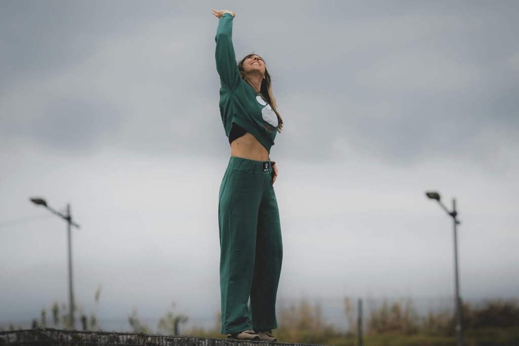 Nainen seisoo vihreässä verkkapuvussa keskellä aukeaa ja kurottelee taivasta. 