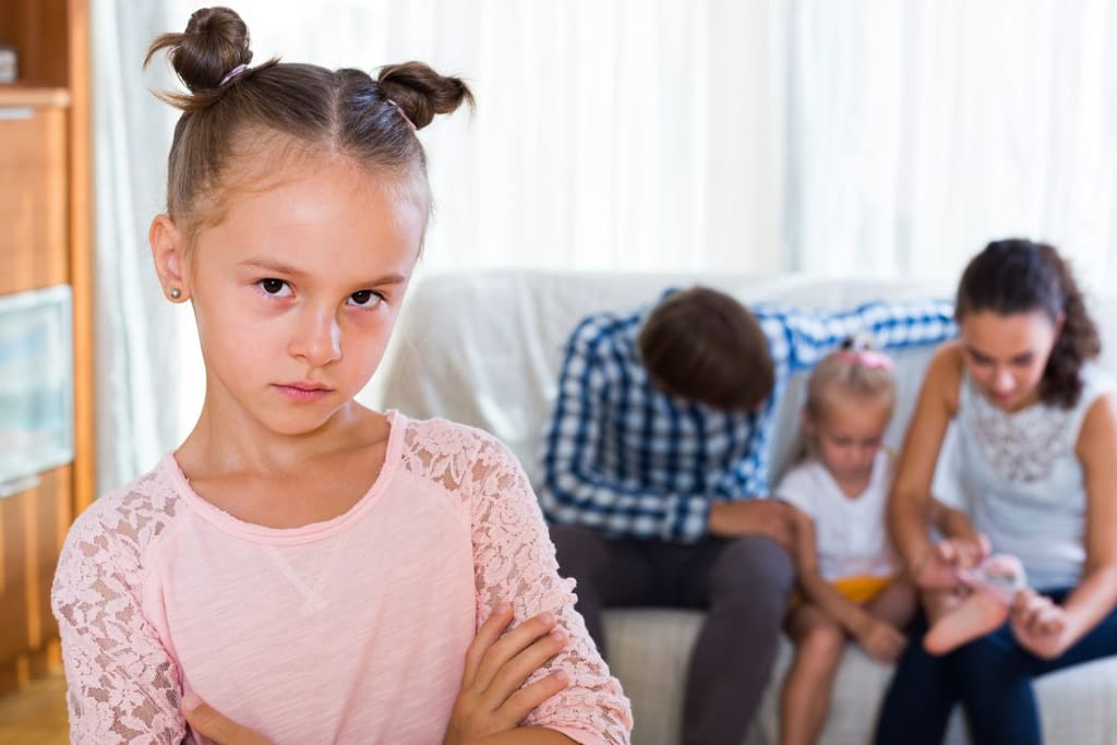 Etualalla lapsi seisoo kädet ristissä ja katsoo kameraan, kun taustalla vanhemmat keskittyvät toiseen lapseen.